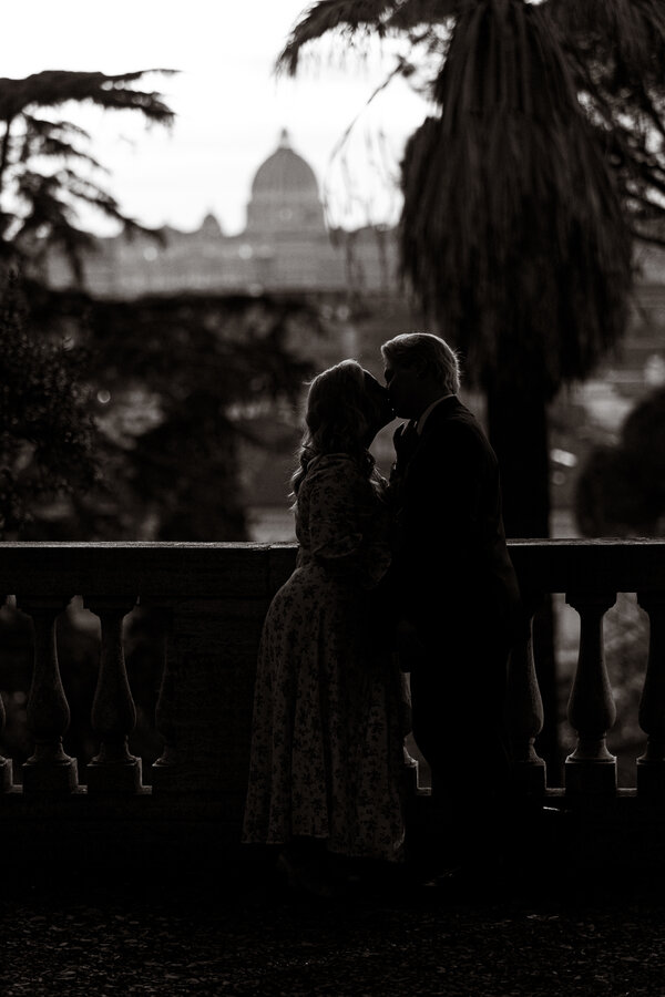 Black and white picture of newly-engaged couple kissing with Saint Peter's dome in the background