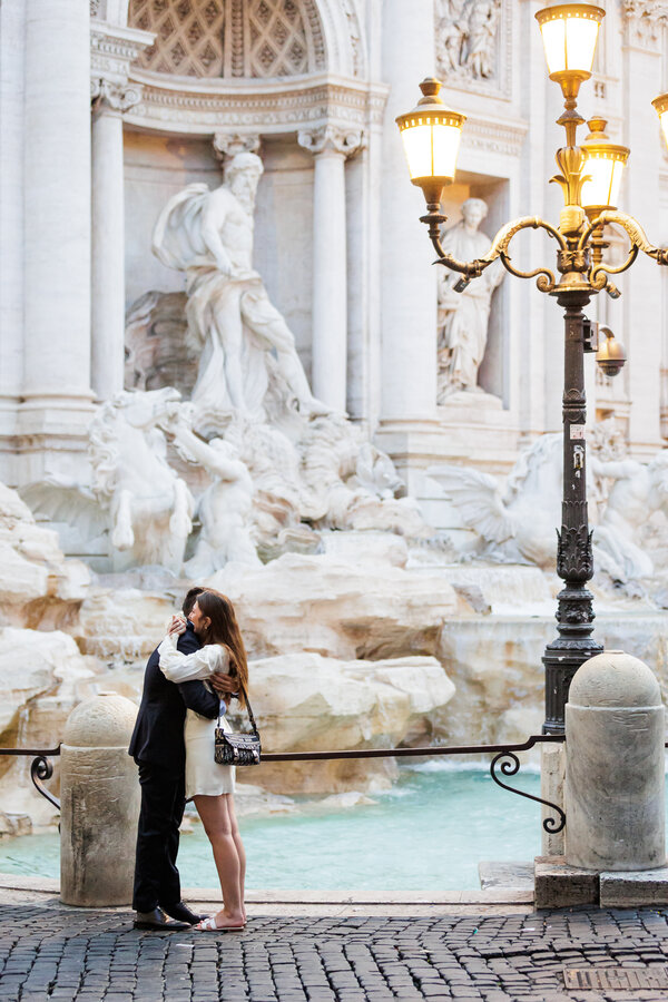 Newly-engaged couple hodling each other during their suprise marriage proposal at the Trevi Fountain at sunrise