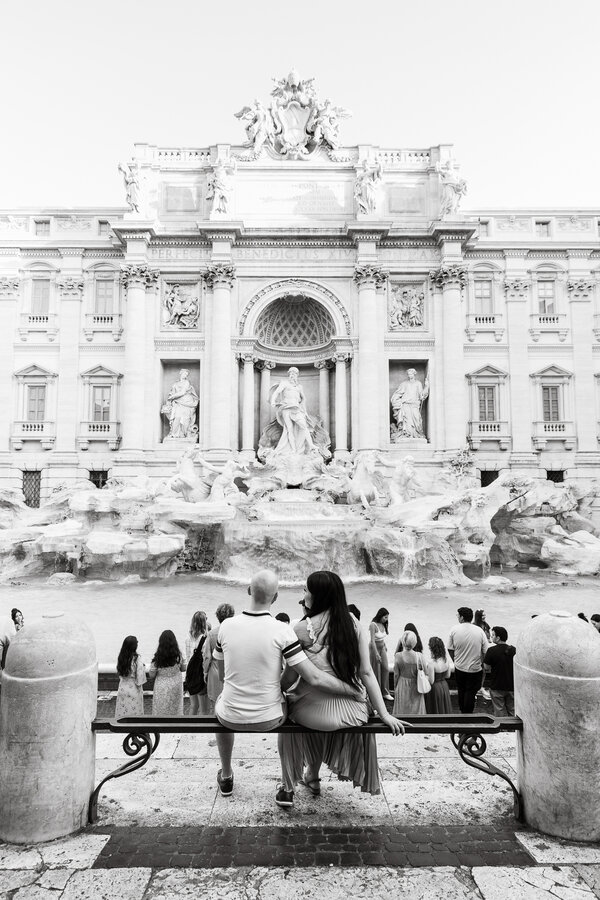 Newly-engaged couple sitting while facing the Trevi Fountain during their engagement photo shoot in Rome