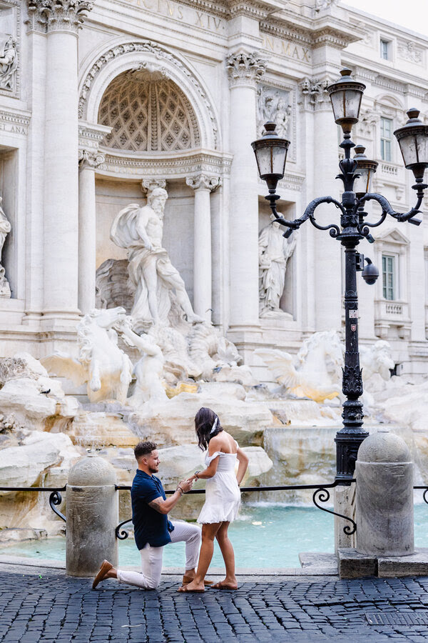 Guy slipping the engagement ring onto his girlfriend's finger during their surprise marriage proposal at the Trevi Fountain