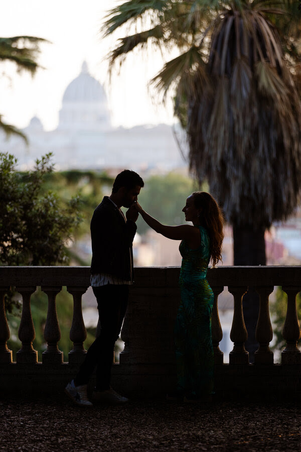 Silhouette of newly-engaged couple with the Vatican in the background while a gentleman kisses his fiancée's hands
