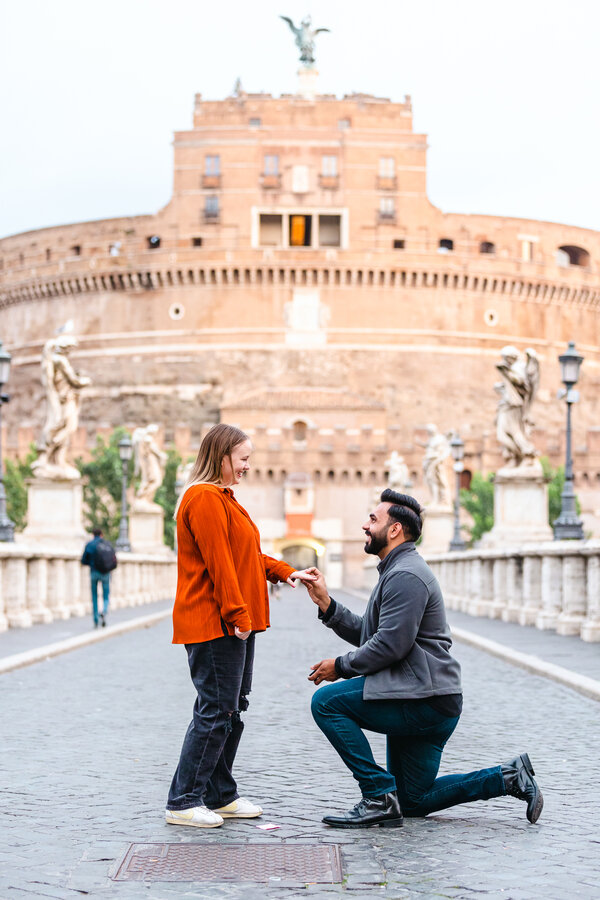 Fiancé kneeling down and putting on the engagement ring on his fiancée on Castel Sant'Angelo Bridge in Rome