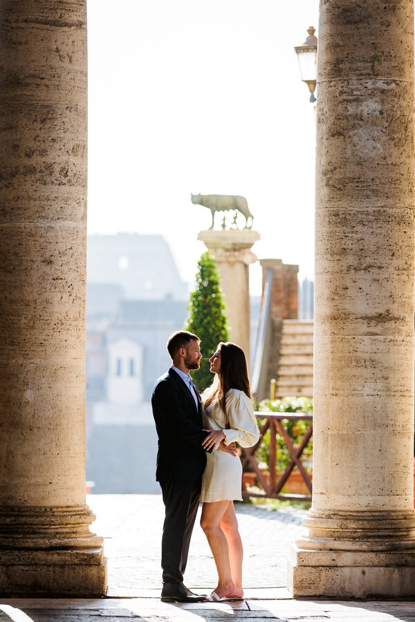 Newly-engaged couple holding each other under colonnade with the she-wolf statue in the background ont heir engagement photo session in Rome