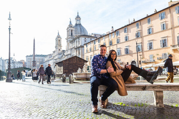 Newly-engaged couple sitting on a bench in Piazza Navona during their secret proposal photo session in Rome