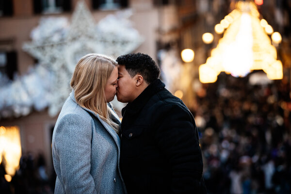 Newly-engaged couple passionately kissing on the Spanish Steps with Via del Corso in the background lit with Christmas lights