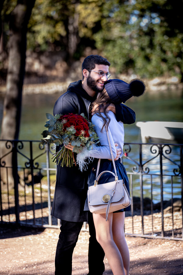 Happy newly-engaged couple with a beauiful bouquet of red roses by the pond in Villa Borghese