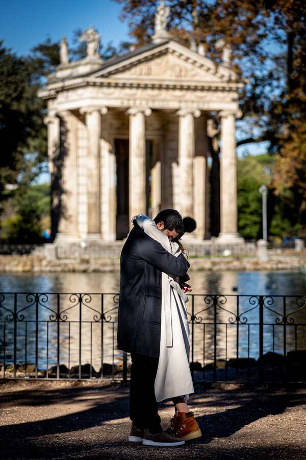Newly-engaged couple holding each other by the pond in Villa Borghese with the Aesculapius Temple in the background