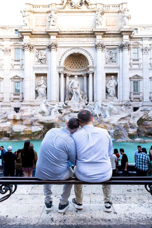 Romantic newly-engaged gay couple sitting next to each other looking at the Trevi Fountain in Rome