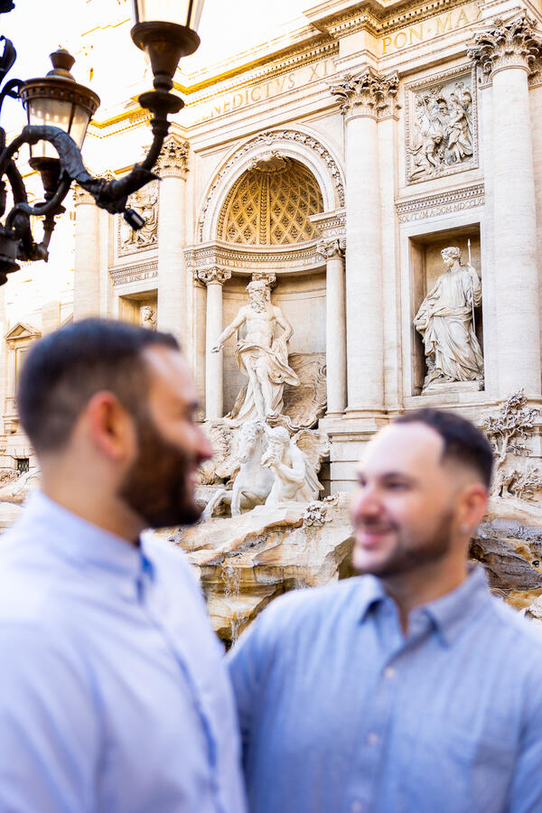 Newly-engaged happy couple looking at each other during their same-sex surprise proposal photoshoot by the Trevi Fountain in Rome
