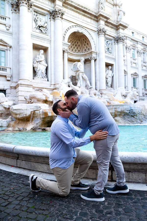 Newly-engaged gay couple kissing during their surprise wedding proposal photoshoot by the Trevi Fountain in Rome