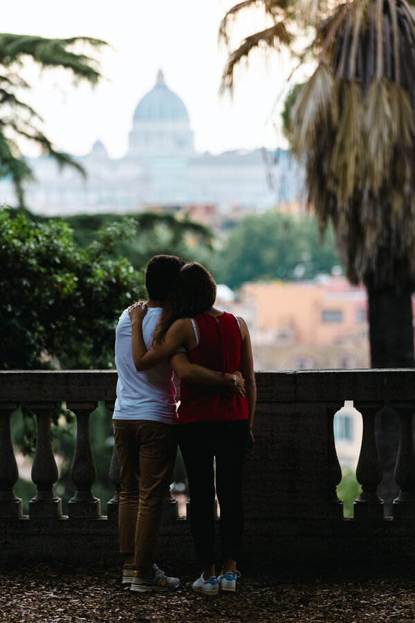 Happy newly-engaged couple holding each other in the Pincio Gardens under the trees with the Vatican in the background