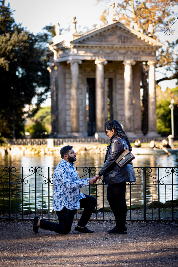 Man kneeling down during his surprise wedding proposal to his girlfriend by the lake in Villa Borghese in Rome