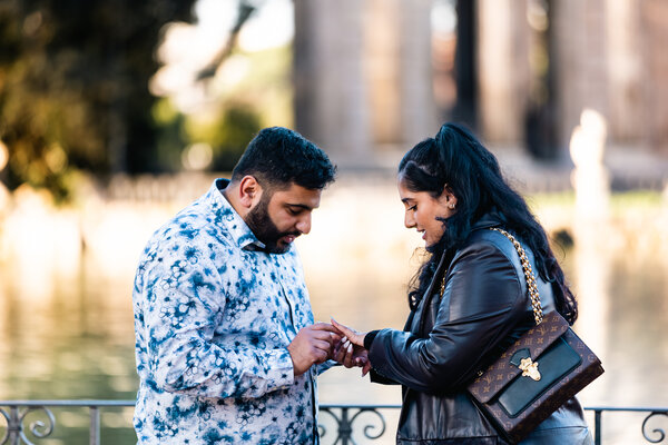 Fiancé fitting the engagement ring on his wife to-be during their surprise marriage proposal by the lake in Villa Borghese in Rome