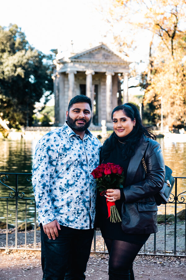 Newly-engaged couple with a red rose bouquet posing for camera during their marriage proposal photo session in Villa Borghese in Rome