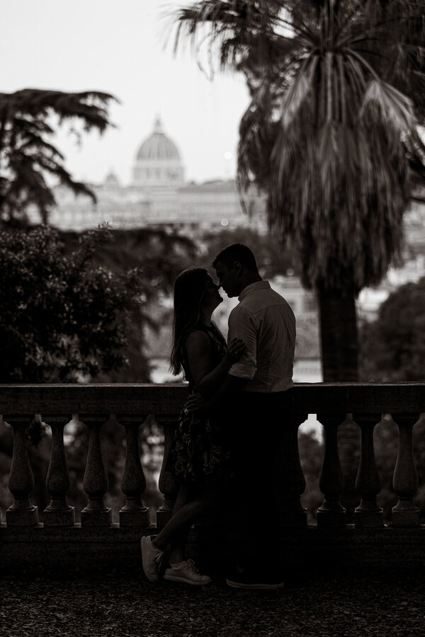 Black and white image of newly-engaged couple in the Pincio Gardens with the Vatican in the background during their surprise proposal photoshoot in Rome