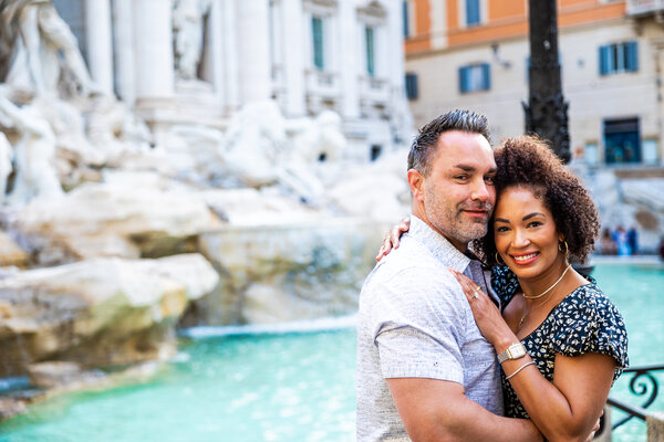 Happy newly-engaged couple at the Trevi Fountain looking into the camera during their surprise proposal photoshoot in Rome