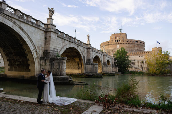 Newly-wed couple holding each other on the Tiber riverbank with Castel Sant'Angelo in the background during their Sposi Novelli photoshoot in Rome