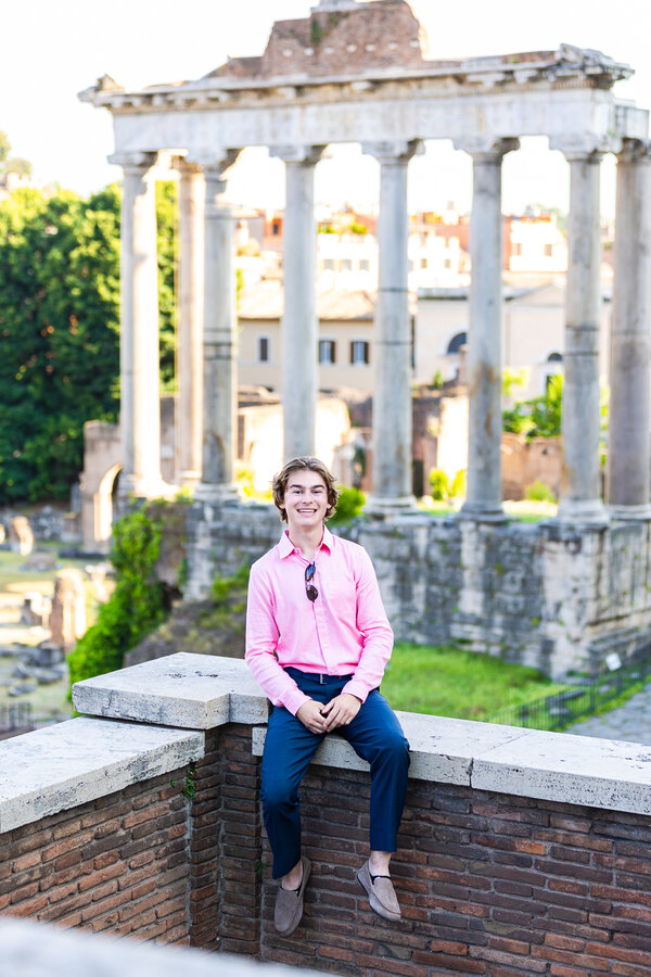 Senior boy sitting on a wall with the Temple of Saturn in the background during his senior photoshoot in Rome