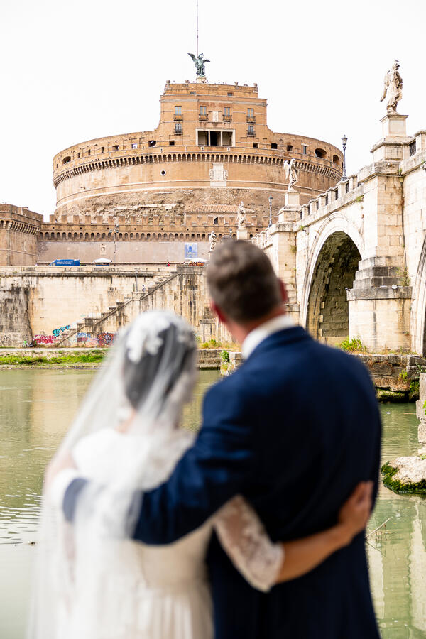 Sposi Novelli couple holding each other and looking at Castel Sant'Angelo from the Tiber riverbank in Rome